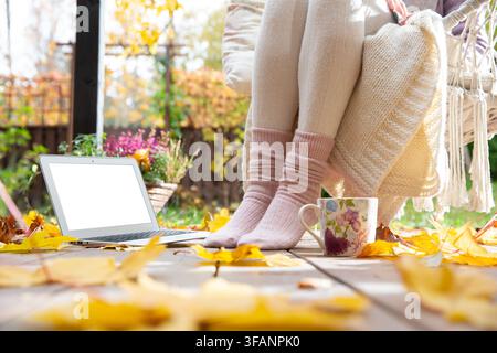 A mid adult woman relaxes on an autumn patio with a laptop mockup, blank screen, and coffee mug - cozy remote work break template concept. Stock Photo