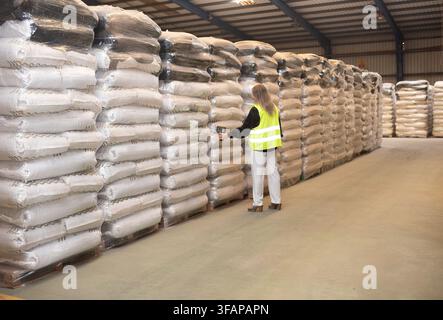 Female warehouse worker in safety vest scans barcodes on stacked bags in storage area Stock Photo