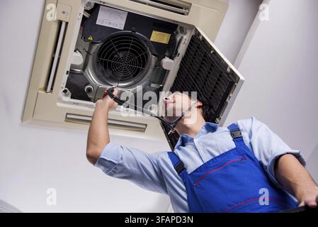 The young mechanic repairs the ceiling air conditioner Stock Photo