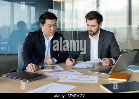Two business partners reviewing and discussing paperwork with charts and graphs on a conference table, demonstrating collaboration and strategy in a modern office environment Stock Photo