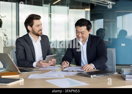 Two diverse business partners collaborating on paperwork, with one man signing a document while the other confidently holding a tablet, discussing financial data in a modern office Stock Photo