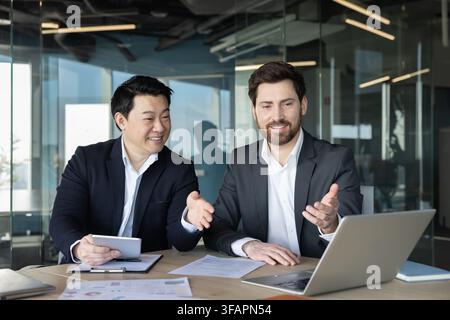 Two diverse male business partners collaborating and discussing a project while reviewing data on a laptop and tablet. Showing teamwork. Partnership. And professional communication in a modern office Stock Photo