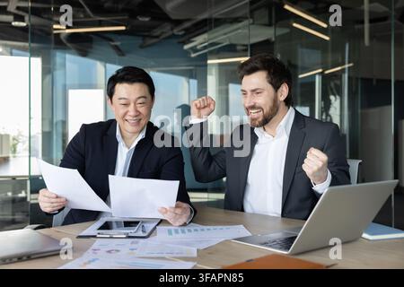 Two cheerful male business partners sitting at a desk in a modern office, one reading documents while the other celebrates a successful deal or project, showing teamwork and partnership Stock Photo