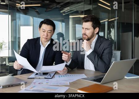 Two diverse businessmen engaging in teamwork, discussing financial reports, and analyzing data during a corporate meeting in a modern office environment Stock Photo