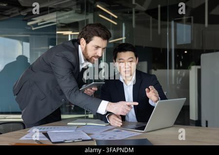 Two diverse business partners collaborating on a new project, working together and analyzing data on a laptop in a modern office meeting, discussing market trends and financial goals Stock Photo