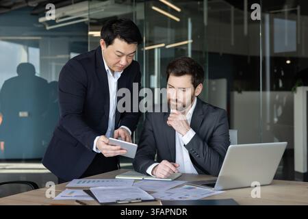 Two diverse businessmen are analyzing data and discussing strategy, collaborating in a modern office environment while utilizing digital technology and paper reports Stock Photo