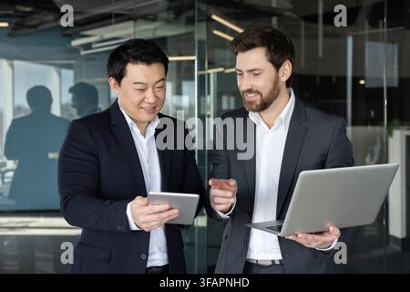 Two diverse businessmen standing in a modern office, analyzing data and discussing business strategies together using a digital tablet and laptop, fostering teamwork and professional development Stock Photo