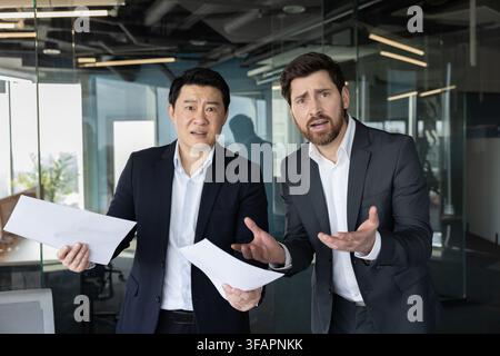 Two diverse businessmen standing in a modern office, holding papers and expressing confusion or misunderstanding, portraying issues like miscommunication, stress, or problem solving Stock Photo