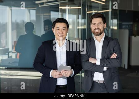 Two confident diverse businessmen, one holding a digital tablet, smiling and standing together in a contemporary office setting, representing successful teamwork and global partnership Stock Photo