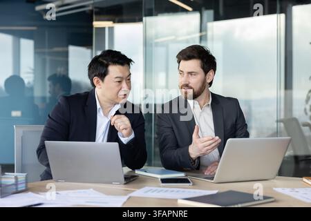 Two male business colleagues of different ethnicities are actively collaborating and brainstorming ideas during a professional discussion in a modern office. Using laptops and documents for their work Stock Photo