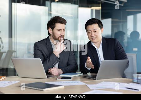 Two diverse business professionals are engaging in a focused discussion and collaborating on a laptop in a modern corporate office, planning strategy and sharing ideas Stock Photo