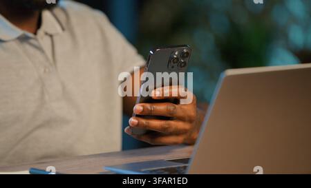 Close up unrecognizable African American business man with laptop using mobile phone app at late night office unknown businessman male guy worker Stock Photo