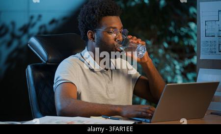 Serious African American architect designer business man typing laptop drinking water in night office focused businessman guy male contractor overtime Stock Photo
