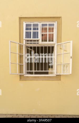 A light yellow wall features a large, white-framed window with multiple panes. The window is open, revealing a barred interior and reflections of a br Stock Photo