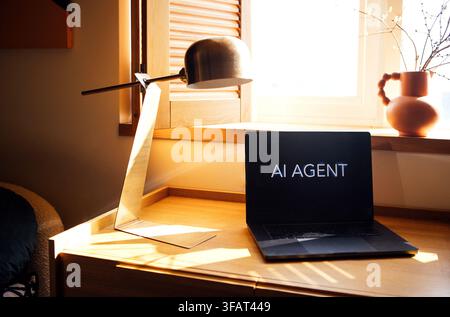 Laptop with "AI AGENT" text sits on a wooden desk next to a modern lamp, illuminated by natural light from a nearby window Stock Photo
