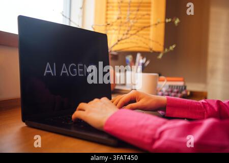 Individual using a laptop with the words AI AGENT visible on the screen, surrounded by a coffee cup and decorative items on a wooden desk Stock Photo