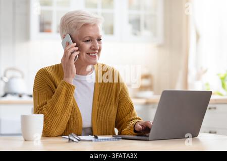 Smiling senior woman having conversation on phone, working on laptop Stock Photo