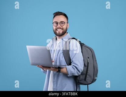 Smart millennial student with backpack using laptop computer over blue studio background Stock Photo