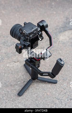 Mirrorless camera mounted on a 3-axis motorized gimbal stabilizer with a tabletop mini tripod on an asphalt ground surface during filmmaking Stock Photo