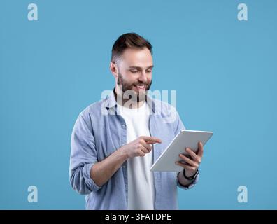 Joyful bearded man using tablet computer for online work or studies on blue studio background Stock Photo