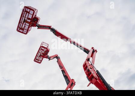 Red articulated boom lifts and hydraulic aerial work platforms extended upward against an overcast sky for industrial building maintenance Stock Photo