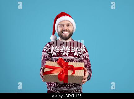 Happy young man in warm sweater and Santa hat holding Christmas gift box on blue studio background Stock Photo
