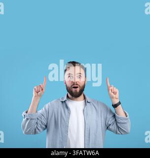 Handsome bearded guy having shocked face expression, pointing at blank space over his head on blue studio background Stock Photo