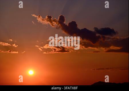 A horizontal photograph capturing a warm golden sunset with the glowing sun descending near the horizon, framed by dramatic dark clouds and faint sunbeams spreading across the evening sky. Stock Photo
