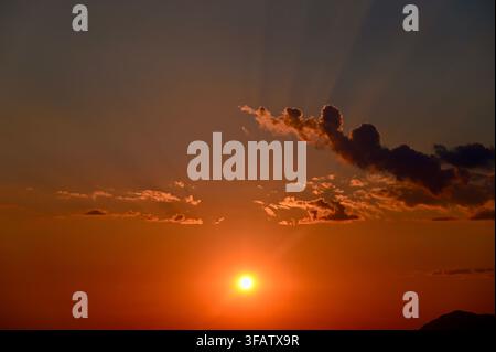 A horizontal photograph capturing a warm golden sunset with the glowing sun descending near the horizon, framed by dramatic dark clouds and faint sunbeams spreading across the evening sky. Stock Photo