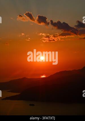 A vertical photograph featuring a bright glowing sun descending over dark mountain silhouettes and a calm sea bay, with dramatic clouds in the warm evening sky. Stock Photo