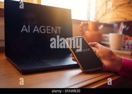 Smartphone shows AI Agent app while being held by a person beside a laptop displaying the same application on a wooden desk with a coffee cup and plan Stock Photo