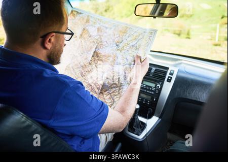 Man tourist sits in driver's seat of car, intently studying large map spread across his lap. With focused expression, traveler carefully planning his route. Stock Photo