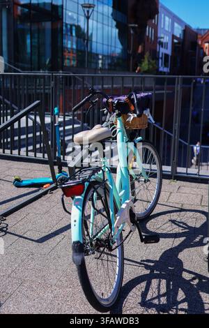 Turquoise city bicycle with wicker basket parked outdoors near modern urban buildings on a sunny day. Stock Photo