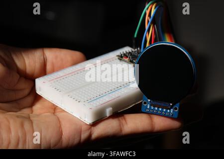 Breadboard connected to microcontroller with jumper cables to round lcd display held in the hand Stock Photo