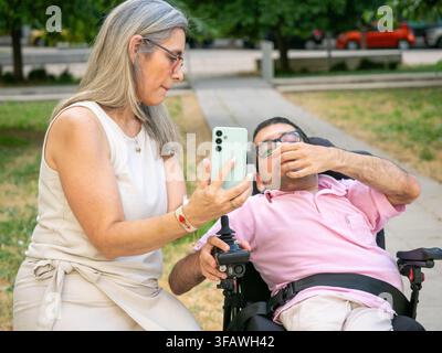 Caregiver assisting man with disability in power wheelchair using smartphone in park Stock Photo