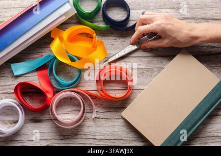 Crafting colorful ribbons, wrapping paper rolls, a gift book and a hand holding scissors on wooden table. Stock Photo