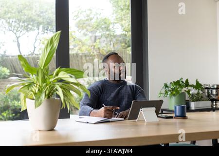 African man sitting at wooden table in home, writing in notebook and using tablet Stock Photo