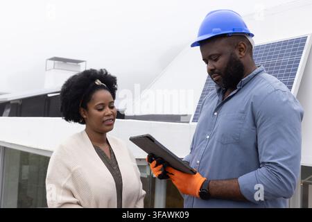 Two African adults standing on rooftop near solar panel, technician in blue hard hat using tablet. Roof, device, safety, helmet, professional, collabo Stock Photo