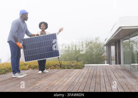 African man and woman installing solar panel on wooden deck with hard hat, gloves, copy space. Terrace, rooftop, misty, photovoltaic, renewable, energ Stock Photo