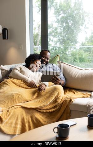 African American couple sitting on sectional sofa, sharing tablet under mustard knit blanket Stock Photo