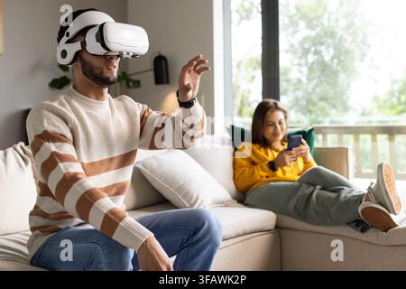 African American couple sitting on sofa, man wearing VR headset gesturing, woman using smartphone Stock Photo