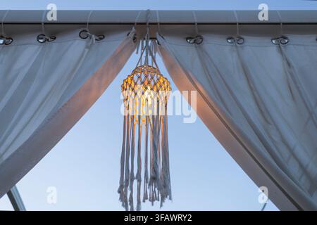 A circular macrame dreamcatcher with long tassels hangs in the center of white curtains. Warm orange light shines from behind the woven fiber decorati Stock Photo