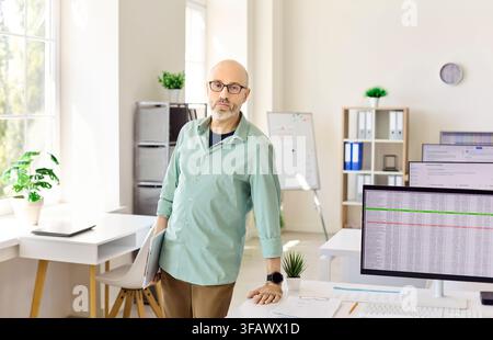 Confident middle aged teacher in modern computer classroom, standing with laptop Stock Photo