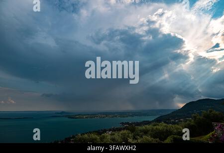 Dramatic storm clouds over lake with sun rays breaking through near hilly shoreline of Garda Lake, Italy Stock Photo