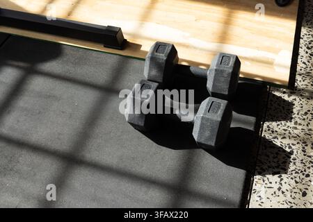 Hex dumbbells resting on black mat near window and wooden lifting platform, shadows crossing. Weights, fitness, minimalist, terrazzo, sunlight, grid, Stock Photo