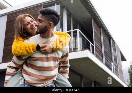 African American couple man giving woman piggyback in yellow sweater in front of home, copy space Stock Photo