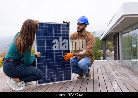 African American team kneeling on deck near sliding door wearing blue helmet installing solar panel. Photovoltaic, outdoor, residential, homeowner, co Stock Photo