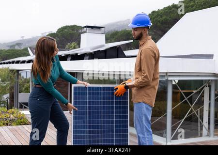 African American coworkers holding and positioning solar panel on rooftop deck with blue hard hat Stock Photo