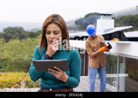 Woman manager holding tablet, reviewing solar on roof while male tech in hard hat handling cables Stock Photo