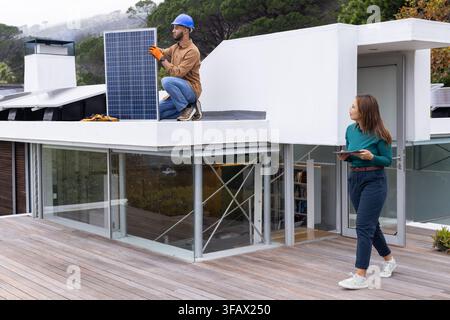 Coworkers installing solar panel on roof man with blue hard hat orange gloves woman holding tablet Stock Photo